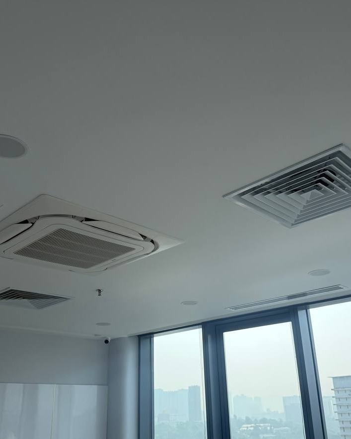 Plaster Ceiling with AC Cassette Fitting – Brickfields Office Flat plaster ceiling in Brickfields office with cassette air-conditioning unit, square diffuser vent, downlights and recessed smoke detector, floor-to-ceiling windows overlooking city skyline