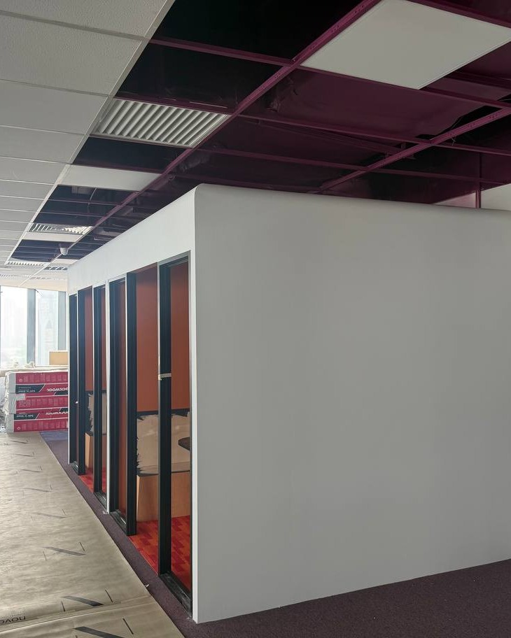 Partition Wall & Ceiling Works – Brickfields Office Freshly installed white partition wall enclosing a soundproof meeting room with black-framed glazed doors and red interior flooring, exposed maroon ceiling grid and suspended ceiling tiles overhead, Brickfields office fit-out