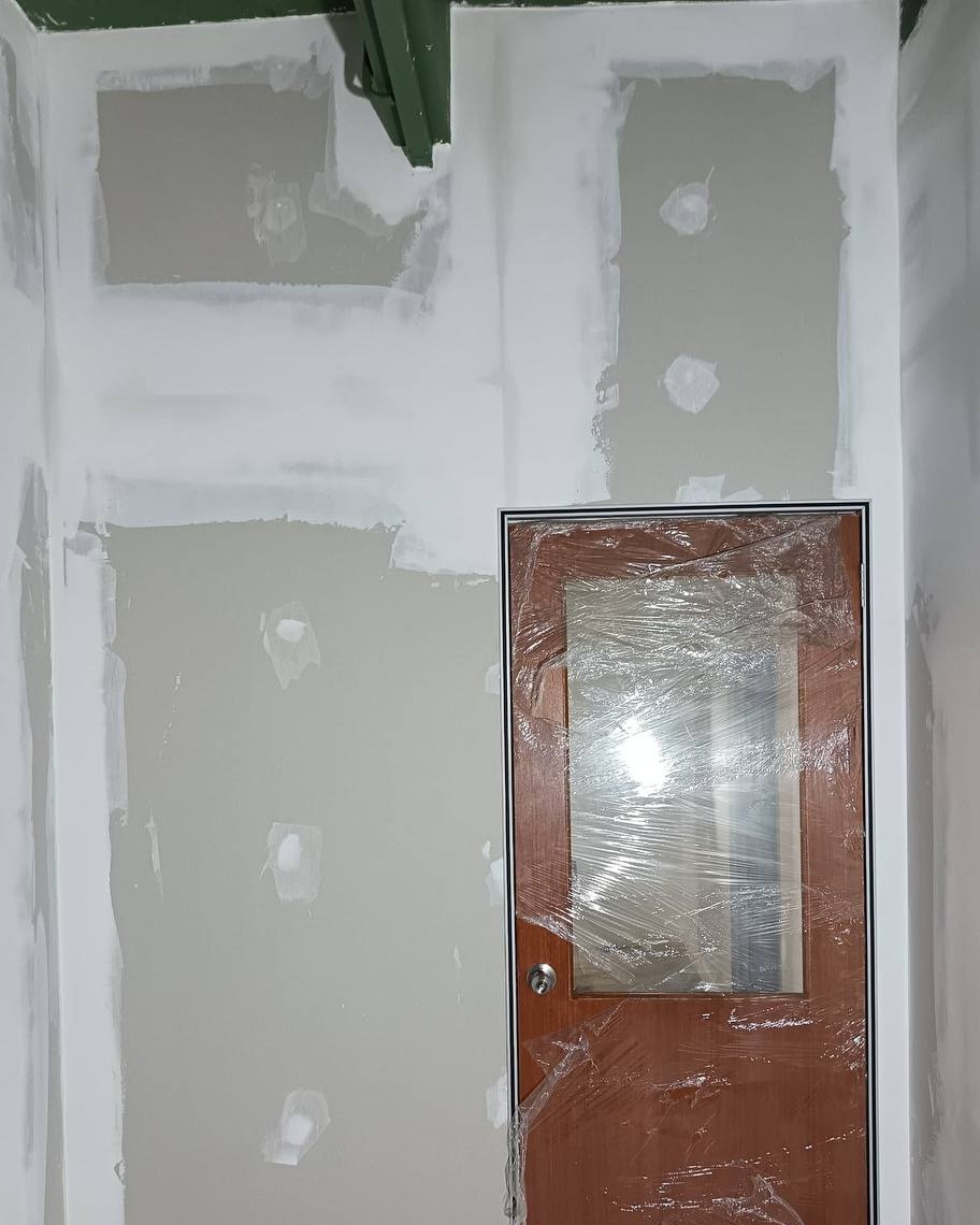 Partition Wall Jointing Near Glass Door – Shah Alam Office Skim-coated plasterboard partition wall with jointing compound patches around a wooden door fitted with glass panel and protective plastic wrap, Shah Alam office partition installation
