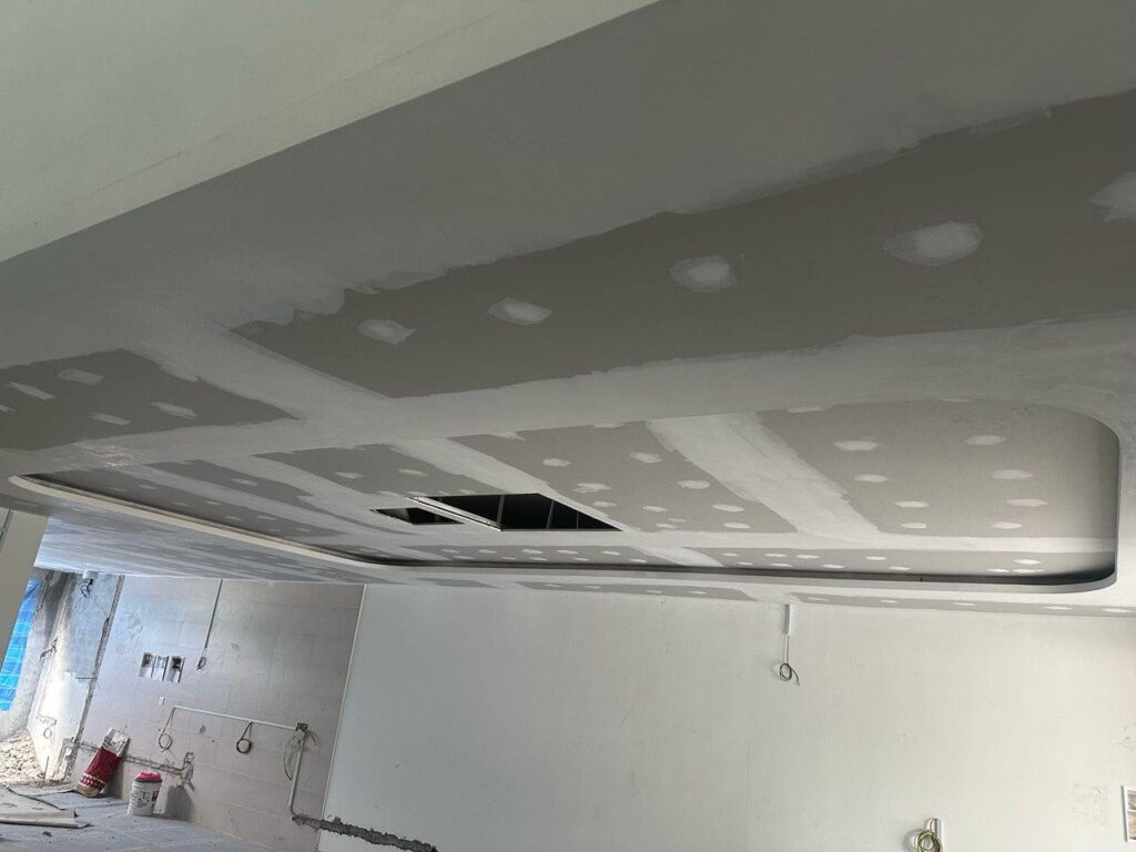 Ceiling construction featuring a curved recessed bulkhead. Joint lines and screw holes are patched, indicating progress before final finishing. Wiring and surface work are still underway.