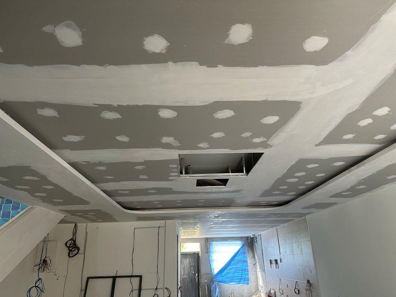Ongoing plasterboard ceiling installation. Joint lines and screw spots are patched with compound, while electrical wiring and ceiling access panels are in progress.