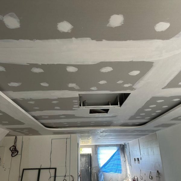 Ongoing plasterboard ceiling installation. Joint lines and screw spots are patched with compound, while electrical wiring and ceiling access panels are in progress.