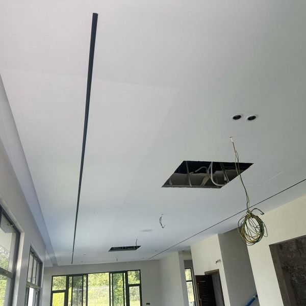 Work-in-progress plaster ceiling featuring a sleek linear groove, spotlight fittings, and wiring access points. Natural light fills the room, highlighting the modern layout.